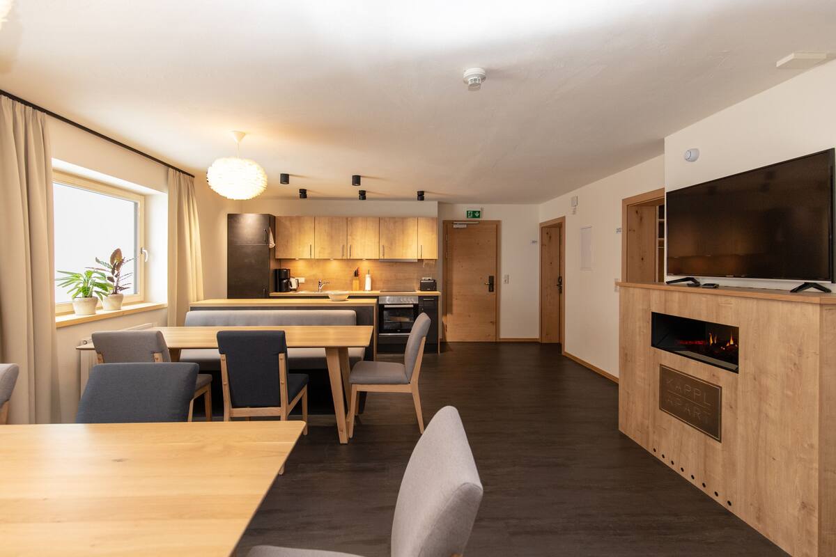 A modern kitchen with wooden cabinets, a dining area with a table and chairs, and a flat-screen TV mounted on a wooden cabinet.