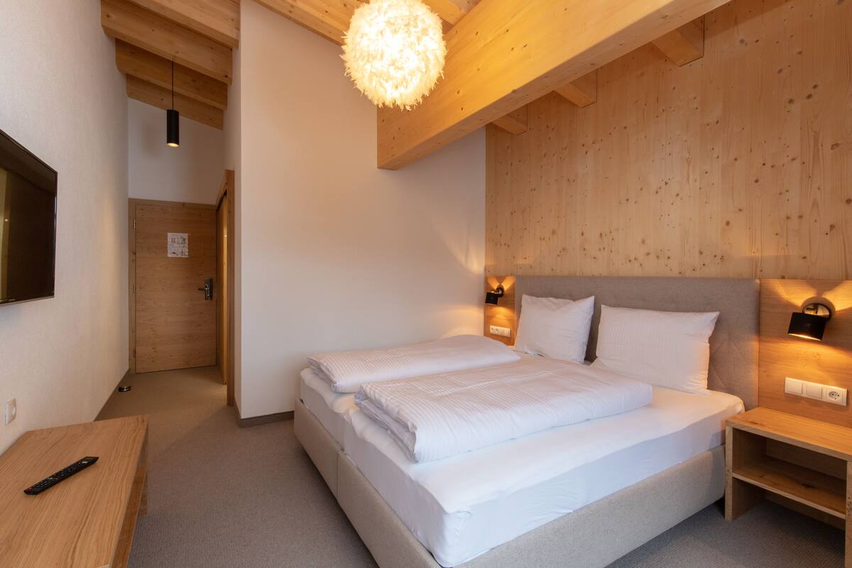 A bedroom with a large bed, wooden ceiling beams, a wooden wall, and a small bedside table.