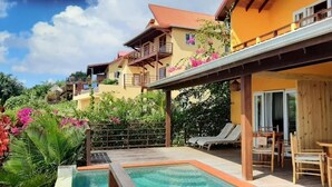 Zona junto a la piscina con sillones de descanso, una pérgola de madera y vistas a un edificio amarillo con balcones.