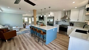 A modern kitchen with white cabinets, a central island with bar stools, stainless steel appliances, and a large window. - The Lazy Tiki - Private Pool (Fort Myers Beach)