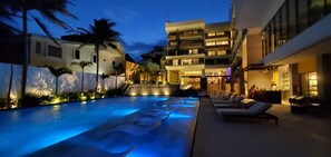 A swimming pool with lounge chairs and a bar area, surrounded by buildings with balconies and a palm tree.