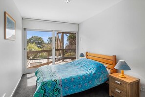 Quarto com cama, mesa de cabeceira, abajur e porta de vidro deslizante que dá para uma varanda.