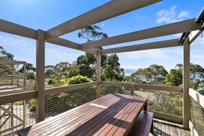 A wooden deck with a table and benches, surrounded by a metal railing, overlooking a scenic landscape with trees and a view of the sea. - Maddlyn with Ocean Views & Peaceful Treed Retreat (Lorne)