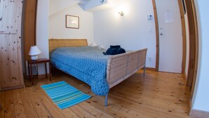 A bedroom with a bed, a bedside table with a lamp, a wooden floor, a door, and a wall-mounted light fixture. - Rent a Room at Cabot Shores - We are on the Cabot Trail (Englishtown)