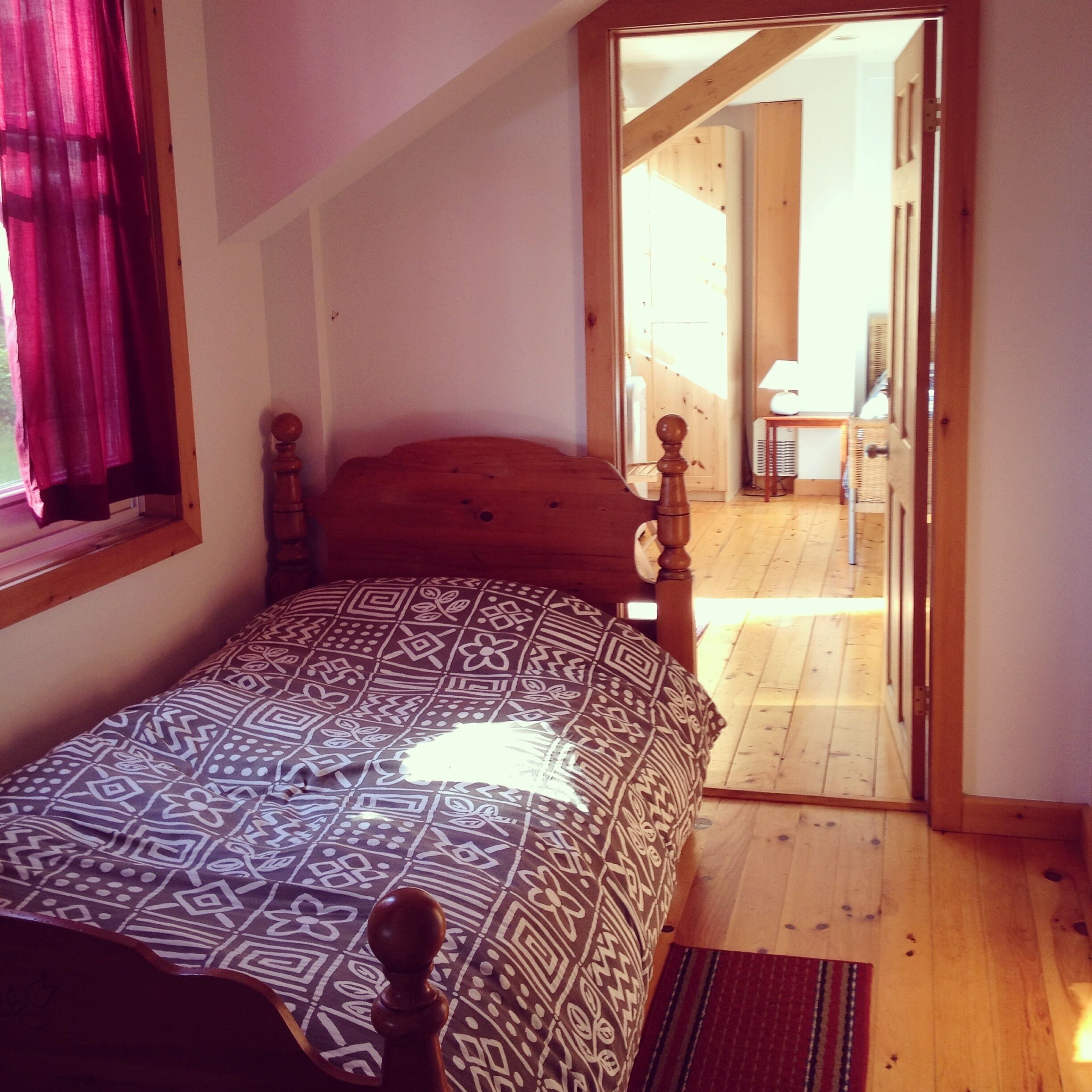 A bedroom with a bed, a window with curtains, a wooden floor, and a door leading to another room.