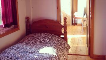 A bedroom with a bed, a window with curtains, a wooden floor, and a door leading to another room.