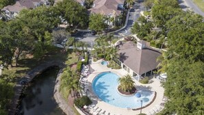 Vue aérienne d’un quartier résidentiel avec une piscine entourée de chaises longues, un petit bâtiment et une route sinueuse.