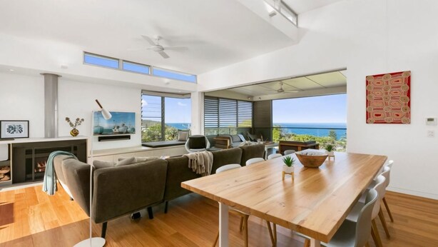 A modern living room with a wooden dining table, comfortable seating, a fireplace, and large windows offering ocean views. - Seaview Tce - Spectacular Home with Stunning Ocean and Headland Views (SUNSHINE BEACH)