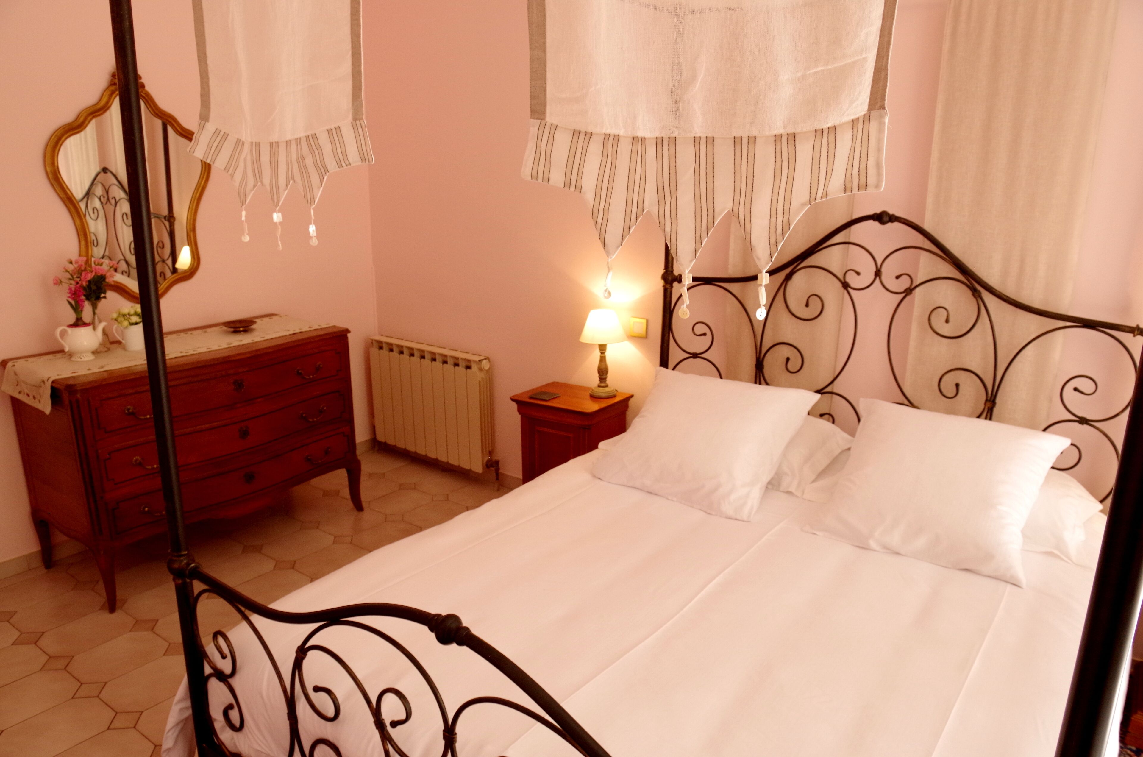 A bedroom with a four-poster bed, a wooden dresser, a nightstand with a lamp, and a mirror on the wall.