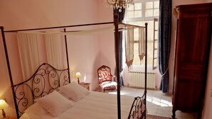 Una cama con dosel adornada con sábanas blancas, una mesita de noche de madera con una lámpara, una silla roja con estampado y un armario de madera.