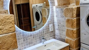 Un baño con pared de piedra, salpicadero de azulejos, lavamanos con espejo y lavadora.