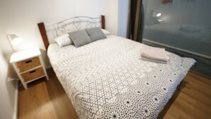 Een net opgemaakt bed met een deken met een geometrisch patroon en twee kussens, een nachtkastje met een lamp, en uitzicht naar buiten door een glazen deur.