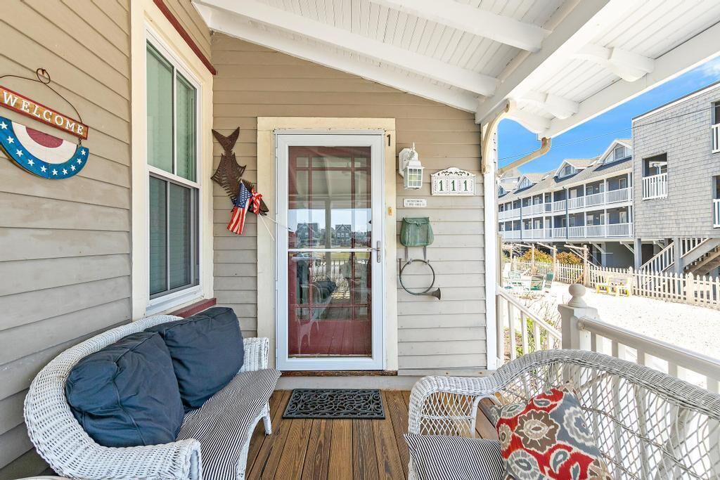 A covered porch with wicker furniture, a welcome sign, and a view of a building with balconies.