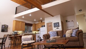 A spacious living area with a wooden ceiling, a dining area with bar stools, a cozy couch, and a kitchen in the background.