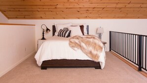 A bedroom with a wooden ceiling, a bed with a white duvet, multiple pillows, a bedside lamp, and a black railing.