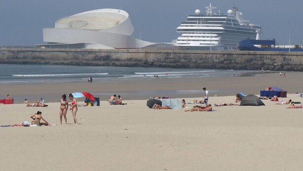 Una playa con gente tomando el sol y nadando, un gran crucero y un edificio blanco distintivo al fondo.