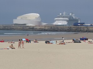 Ein Strand mit badenden und sonnenden Menschen, einem großen Kreuzfahrtschiff und einem markanten weißen Gebäude im Hintergrund.