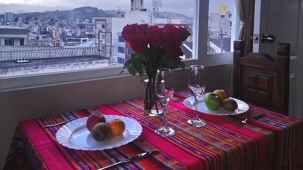 A table set with plates, glasses, and a vase of red flowers, with a cityscape view through a window.