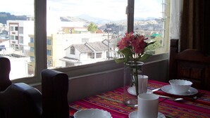 A dining table with white cups and saucers, a vase with pink flowers, and a view of buildings and mountains through a window.