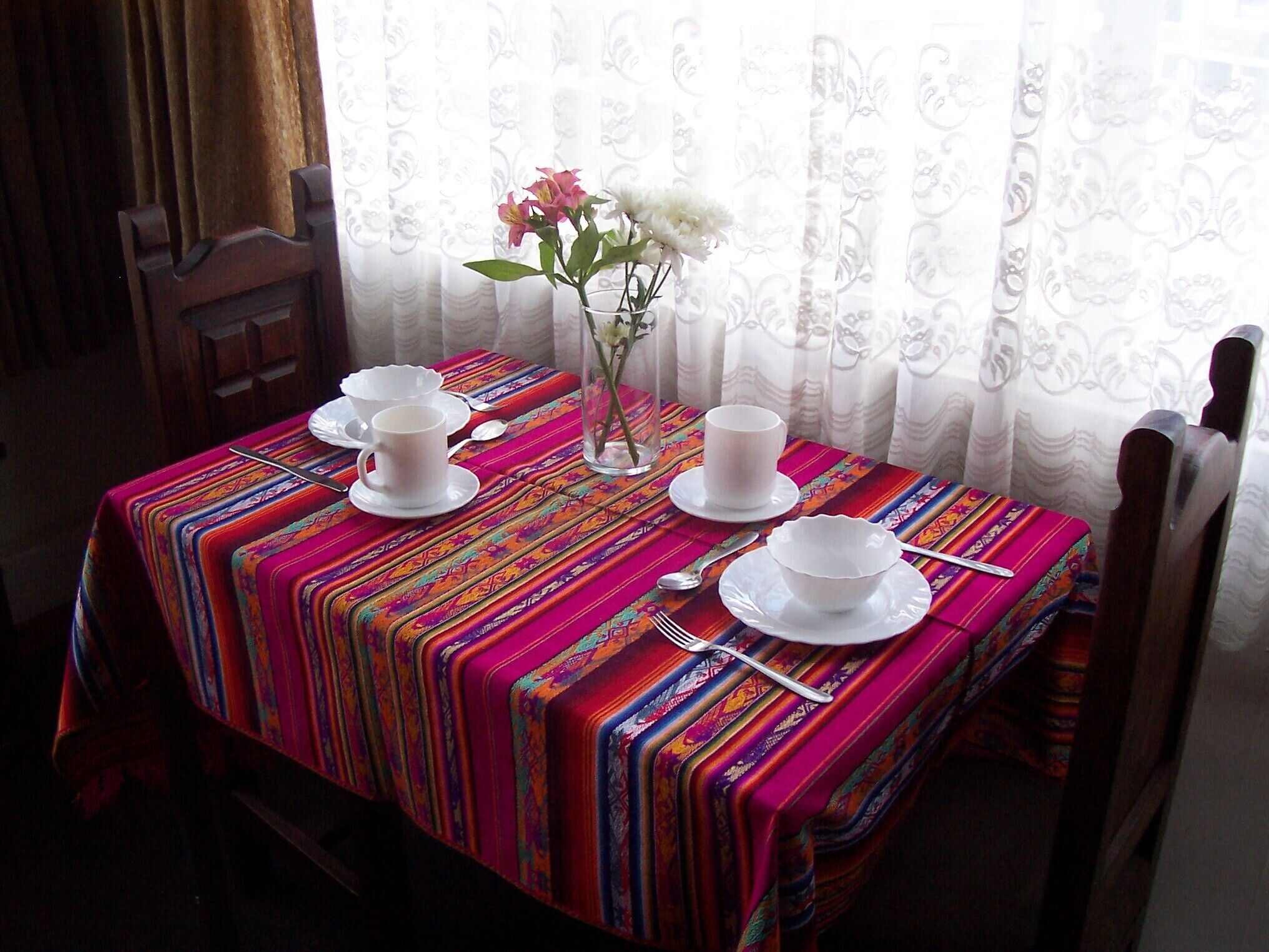 Une table dressée avec des tasses et des soucoupes blanches, un vase rempli de fleurs, le tout recouvert d’une nappe rayée aux couleurs vives.