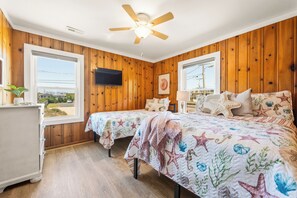 A bedroom with two beds, wooden walls, a ceiling fan, a TV, and a window with a view.