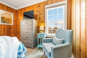 Ein Zimmer mit Holzwänden, einem Fernseher auf einem Schrank, einem Fenster mit Aussicht, einem blauen Sessel und einer Lampe auf einem Beistelltisch.