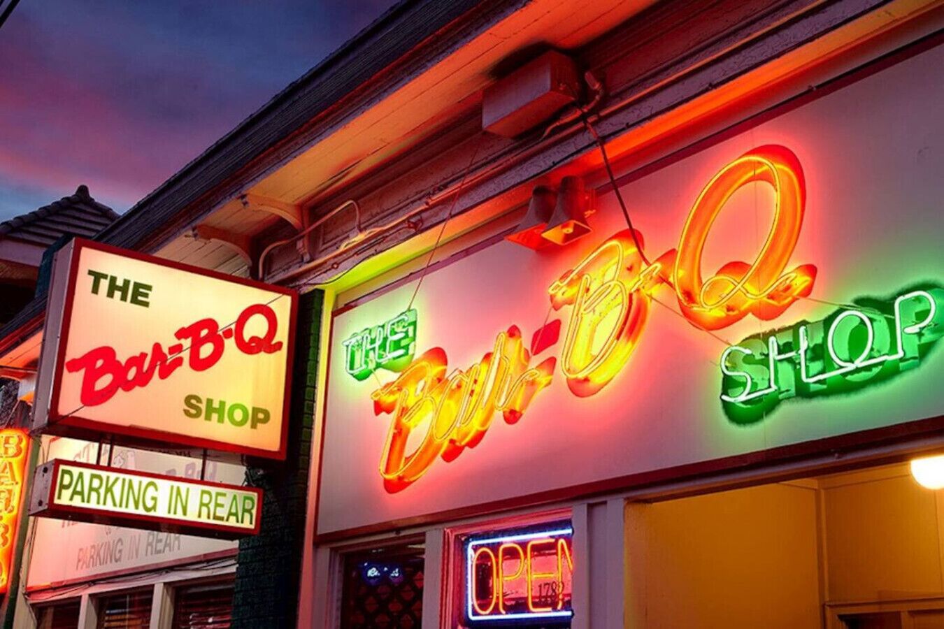 A storefront with neon signs for 'The Bar-B-Q. Shop' and 'The BBQ Shop' along with a 'PARKING IN REAR' sign.