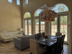 A spacious living room with a large dining table, comfortable seating, and large windows offering views of the outdoors. - Modern Tustin Home  Tesla Universal EV  30+ Days (Tustin)