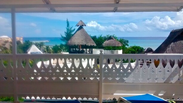 A view from a covered area with a white lattice fence, overlooking a beach with thatched-roof structures and a pier. - Plantation House - Ocean view - Family and pet friendly (Playa del Carmen)