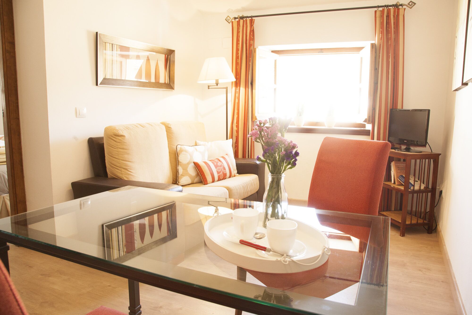 A living room with a glass table, a sofa, a chair, a TV, and a window with curtains.