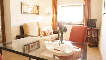 A living room with a glass table, a sofa, a chair, a TV, and a window with curtains.