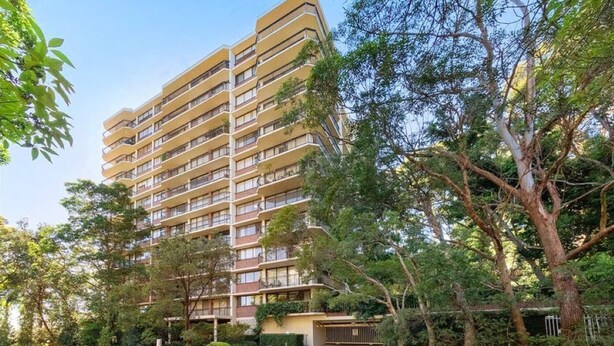 A multi-story residential building with balconies, surrounded by trees and greenery.