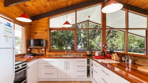 A kitchen with wooden walls, white cabinets, a stove, a microwave, and large windows. - La Tienda (Lorne)