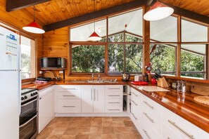 A kitchen with wooden walls, white cabinets, a stove, a microwave, and large windows. - La Tienda (Lorne)