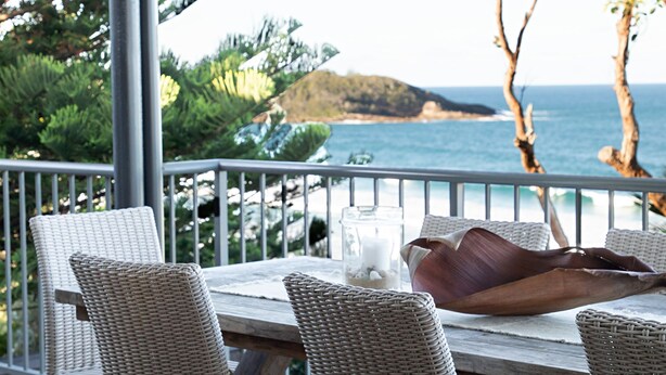 A wooden table with wicker chairs on a balcony overlooking a coastal view with a mountain in the distance. - Kalinda (Mollymook Beach)
