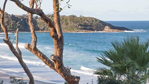 A coastal landscape with a beach, ocean waves, and a forested hill in the background. - Kalinda (Mollymook Beach)