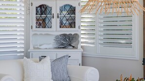 A room with white blinds, a white sofa with patterned cushions, and a decorative shelf with a whale and a branch. - Kalinda (Mollymook Beach)