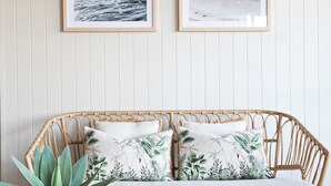 A wicker sofa with two pillows featuring a tropical leaf pattern, set against a white paneled wall with two framed artworks above. - Kalinda (Mollymook Beach)