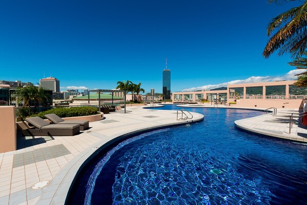 A rooftop pool with lounge chairs, a view of a city skyline, and a clear blue sky.