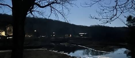 Une vue nocturne d’une rivière où se reflètent les lumières, entourée d’arbres et de bâtiments éloignés.
