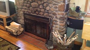 Una chimenea de piedra con un alero adornado con piñas y velas rojas, un televisor sobre un soporte a la izquierda y una cesta de leña a la derecha.