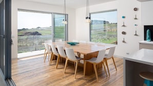 Un espace repas moderne avec une grande table en bois, des chaises blanches et de grandes fenêtres donnant sur un paysage rural.