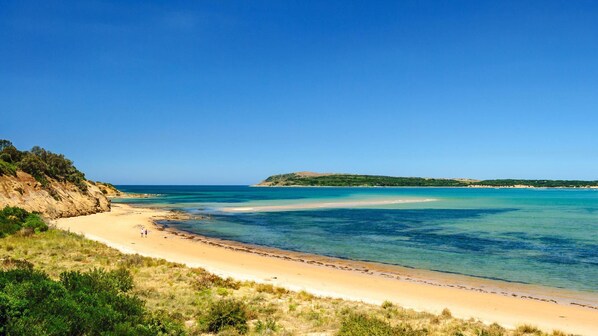 A beach with clear blue water, sandy shore, and a small hill in the distance.