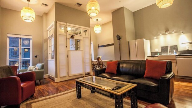 Sala de estar com mesa de centro de vidro, sofá de couro e poltronas vermelhas. Há uma área de cozinha com geladeira e uma mesa de jantar.