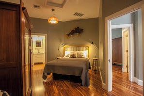 A bedroom with a wooden floor, a bed with a headboard, bedside lamps, and a hanging light fixture.