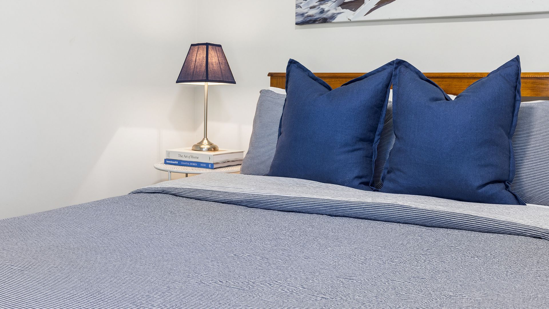 A neatly made bed with a striped bedspread and two pillows, a bedside lamp, and a book titled 'The Art of Home' on a bedside table.