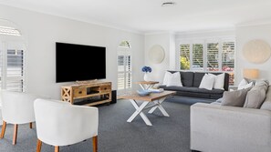 A modern living room with a flat-screen TV, a grey sofa, white armchairs, and a coffee table.