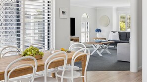 A modern dining area with a wooden table and white chairs, a kitchen in the background, and a grey sofa in the living area.