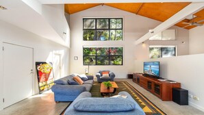 Un salon spacieux avec un haut plafond, de grandes fenêtres, un téléviseur et un coin salon confortable.