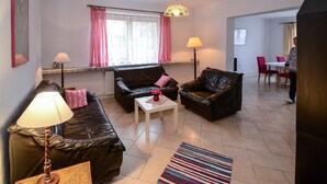 Un salon comprenant un canapé, deux fauteuils, une table basse et une lampe. Une fenêtre ornée de rideaux roses est visible, et on aperçoit un coin repas au fond de la pièce.